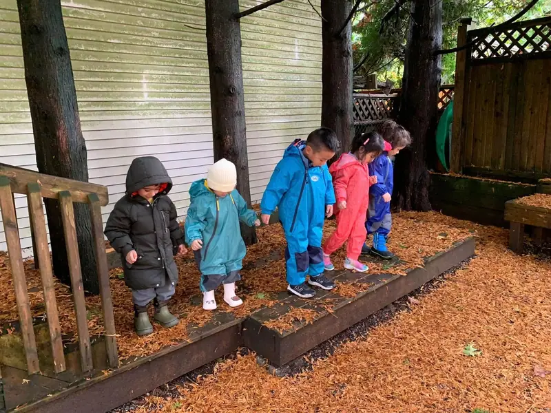 Children in rain suits walking single file along the bark-mulch path in the centre's yard