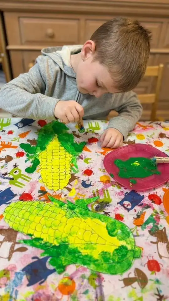 A finished corn-cob painting drying among the paints on the craft table