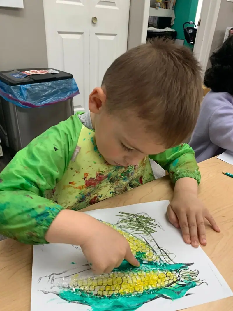 A child in a paint smock drawing a corn cob